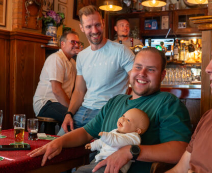 mannen aan tafel waarvan 1 er een neppe baby vast heeft met een wit truitje