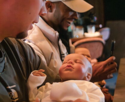 man op dadshower houdt neppe baby vast met witte romper aan, op achtergrond andere man