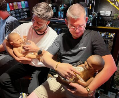 2 mannen op babyshower houden beide nep baby vast en oefenen met fles geven